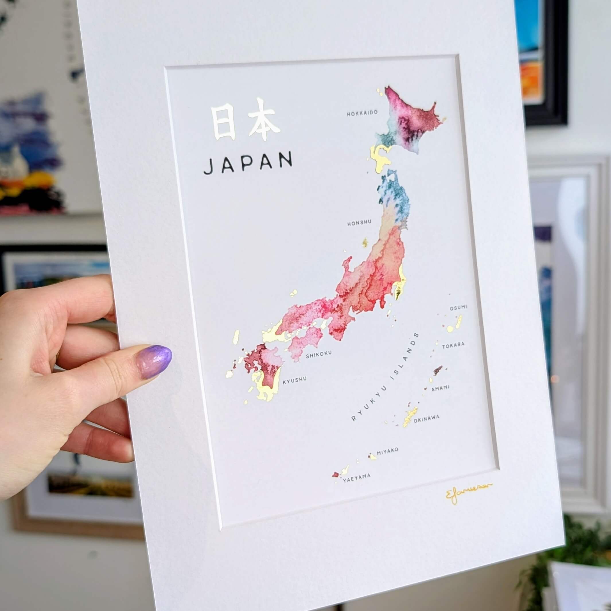 Hand holding a watercolor map print of Japan with gold foil accents, showing regions like Tokyo, Osaka, and Okinawa