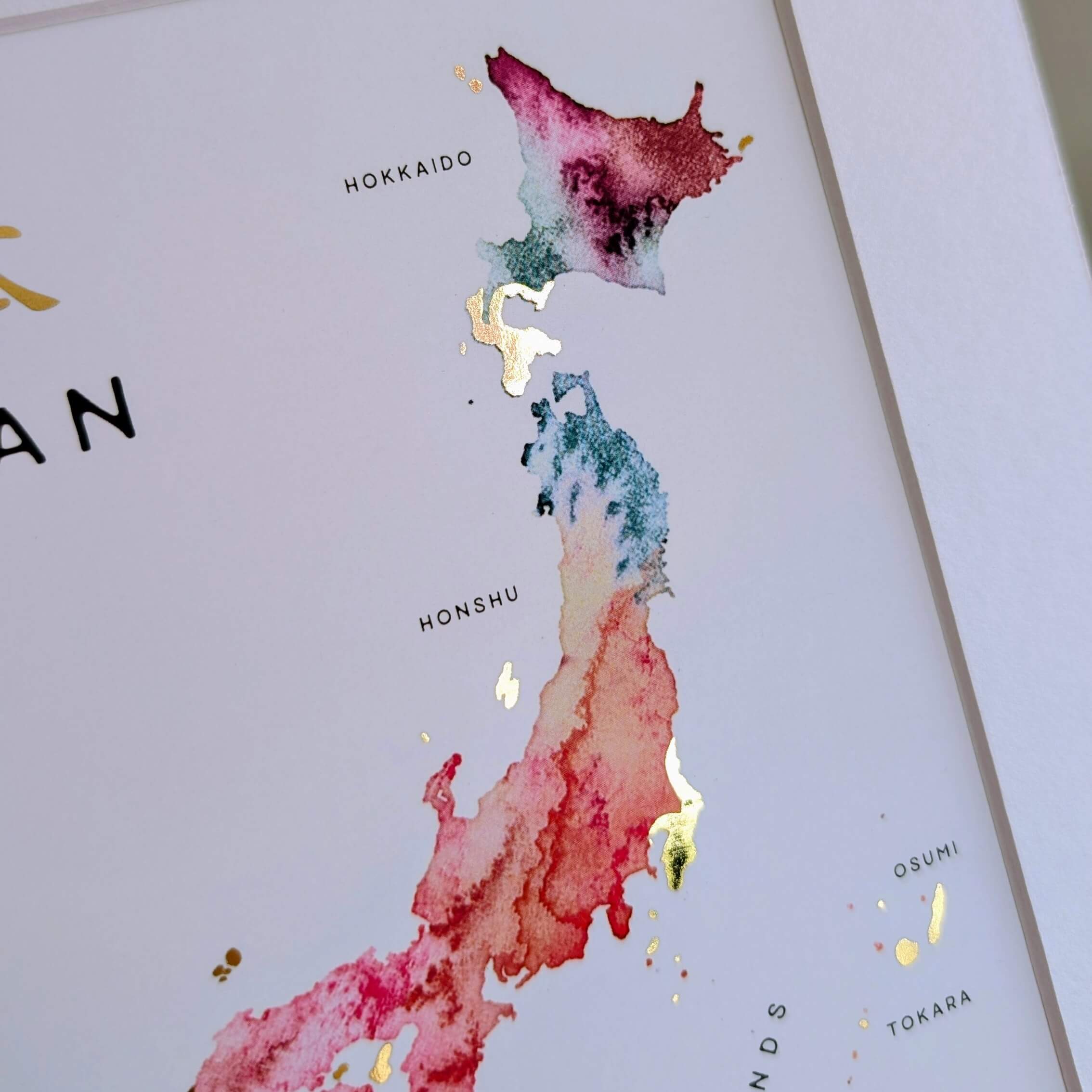Angled shot of a Japan watercolor map print held by hand, showing gold foil shimmer across the islands and subtle texture of the fine art paper