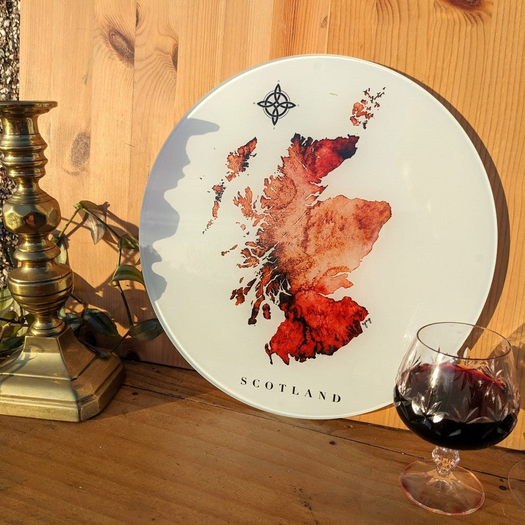 Round decorative plate with a watercolour map of Scotland on a wooden surface with a candle and glass of red wine.